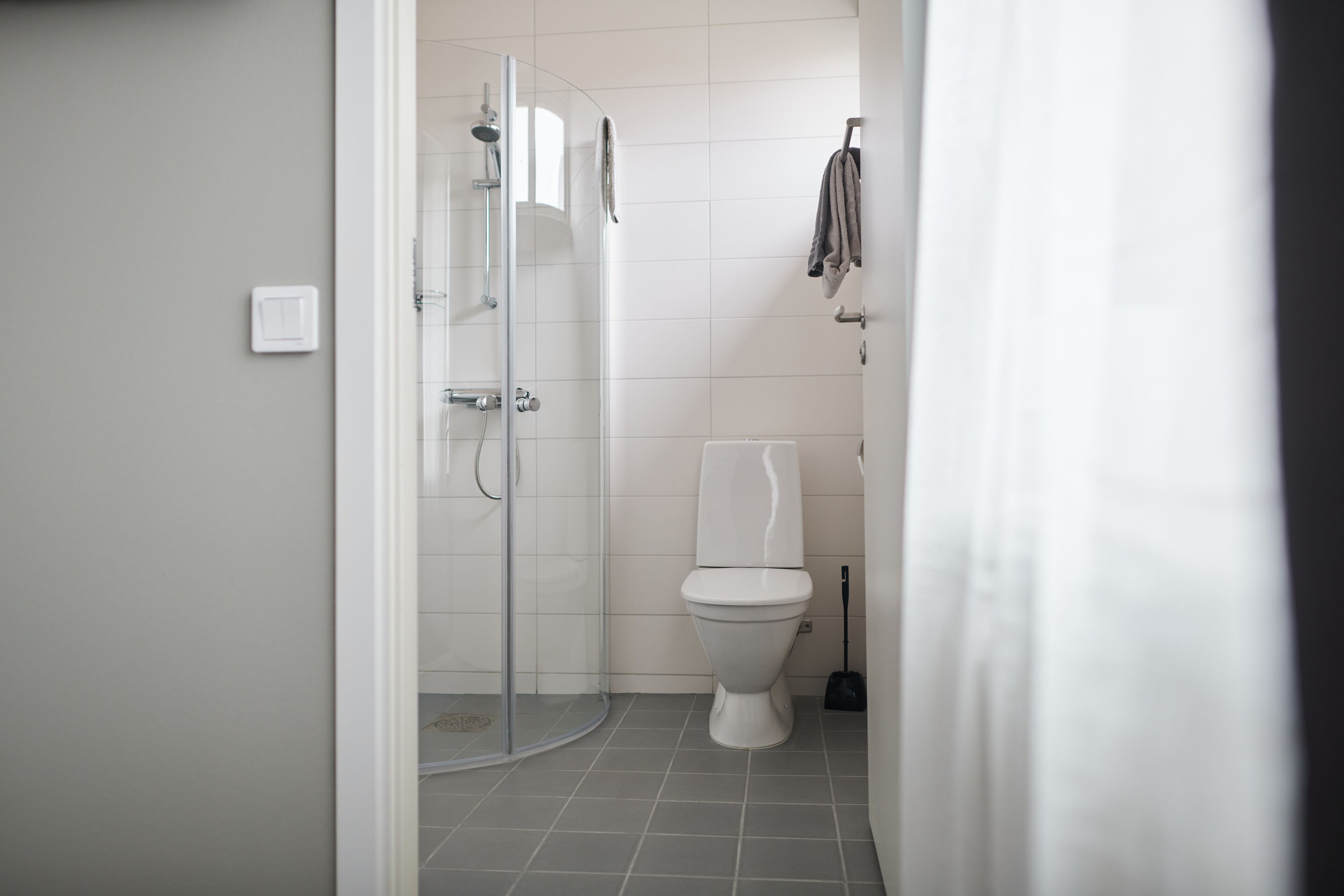 Un baño con cabina de ducha de vidrio, un inodoro blanco y una escoba apoyada en la pared.