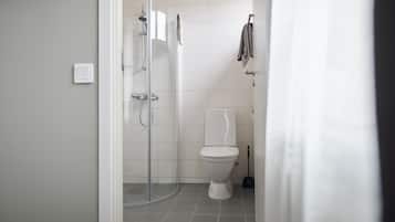 Un baño con cabina de ducha de vidrio, un inodoro blanco y una escoba apoyada en la pared.