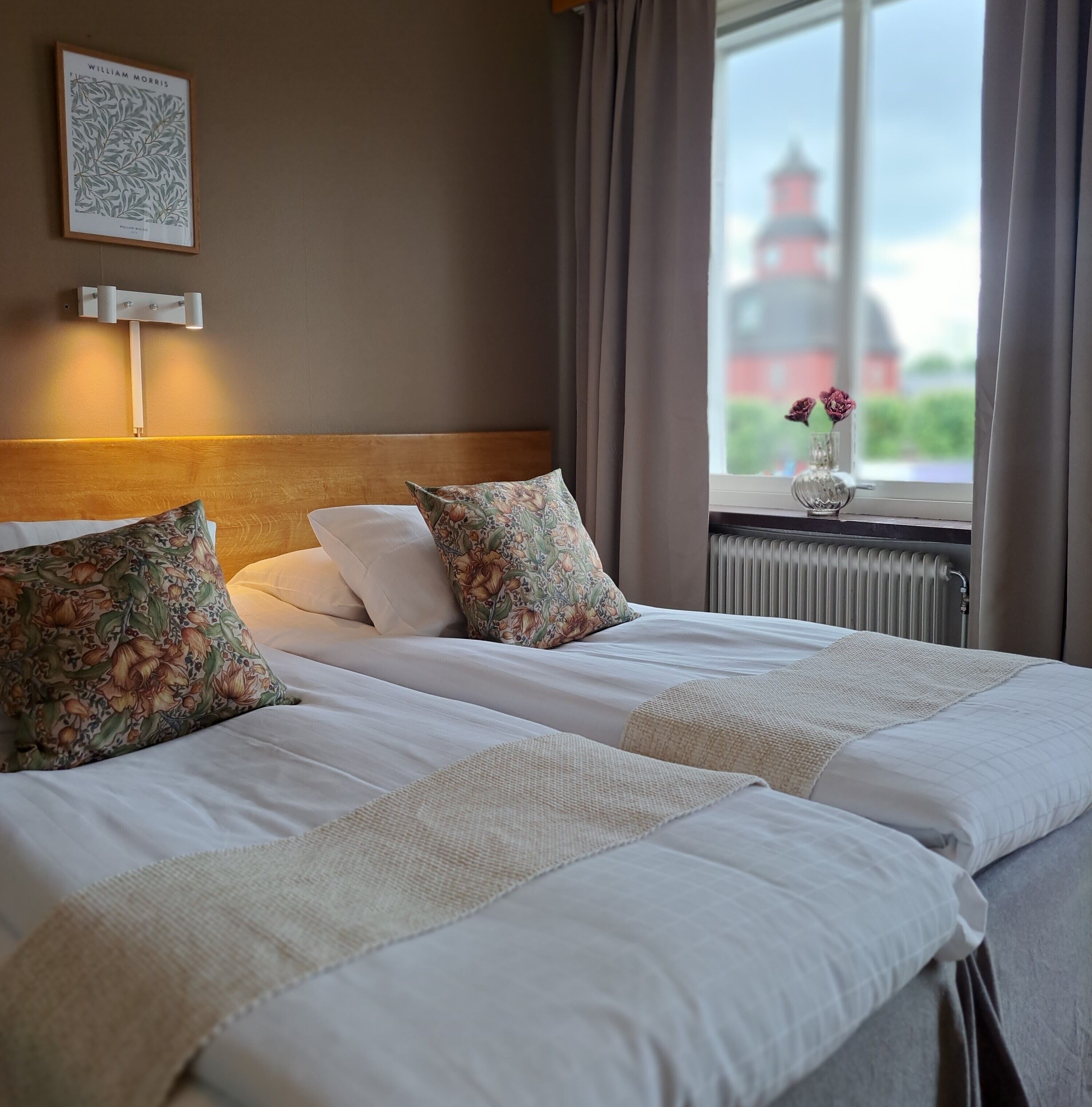 A neatly made twin bed with floral pillows, a framed botanical print on the wall, and a view of a building through a window.