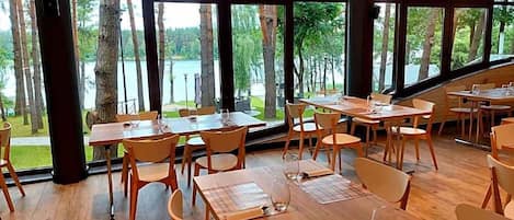 A restaurant with tables and chairs arranged for dining, large windows offering a view of trees and a body of water.