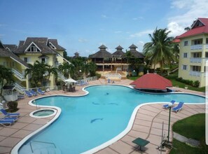 A swimming pool with lounge chairs and a covered area, surrounded by buildings with balconies and palm trees.