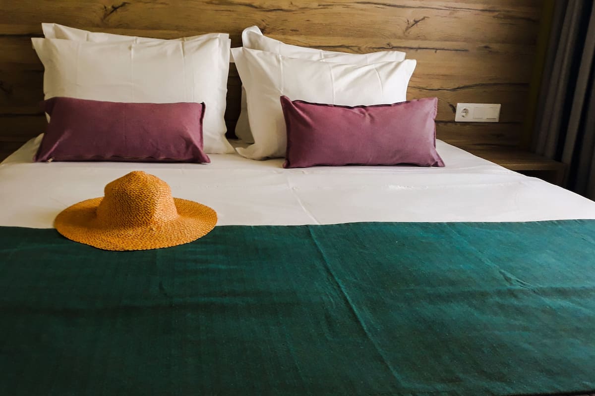 A bed with white and purple pillows, a green and teal bedspread, and a straw hat on the bed.