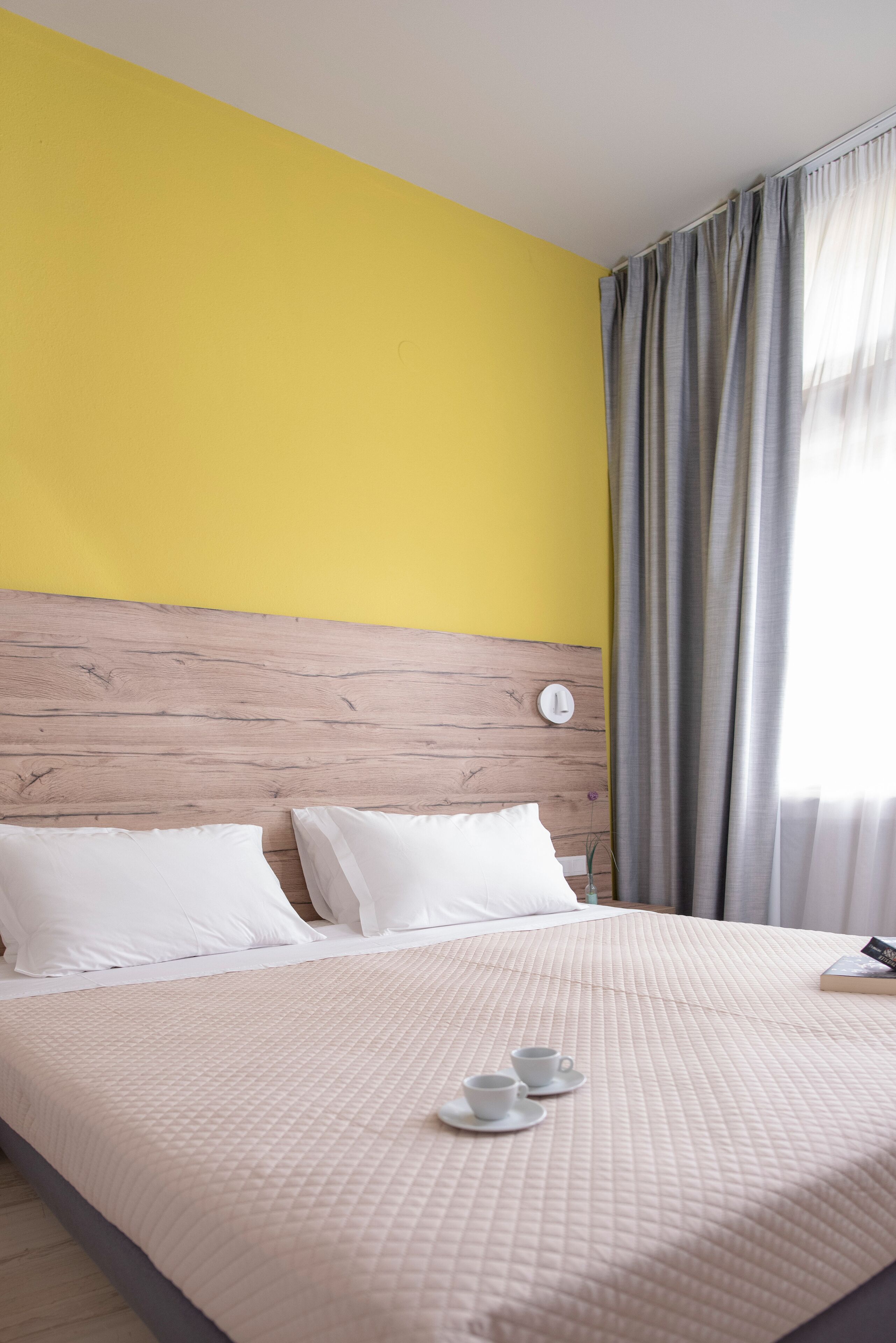 Quarto de hotel com cabeceira de madeira, duas almofadas brancas, cama com colcha bege acolchoada, duas xícaras brancas em pires e um relógio na cabeceira.