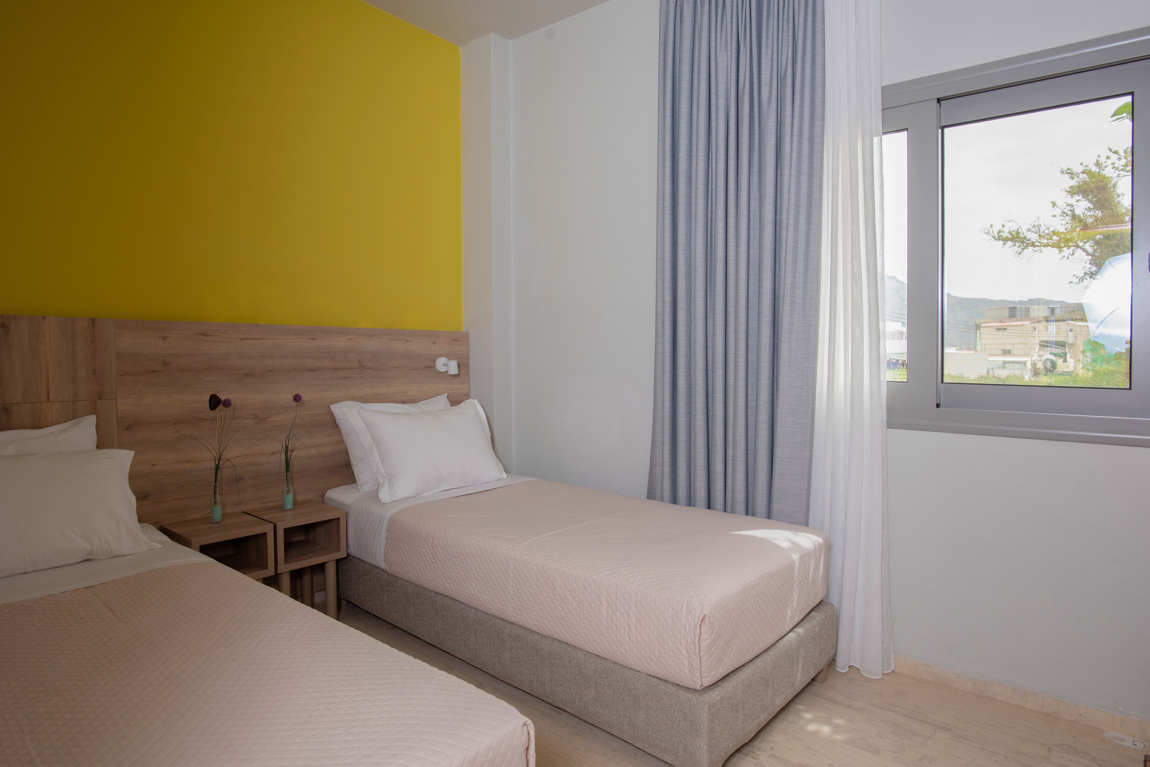 A hotel room with two beds, a wooden headboard, a window with curtains, and a view of buildings outside.
