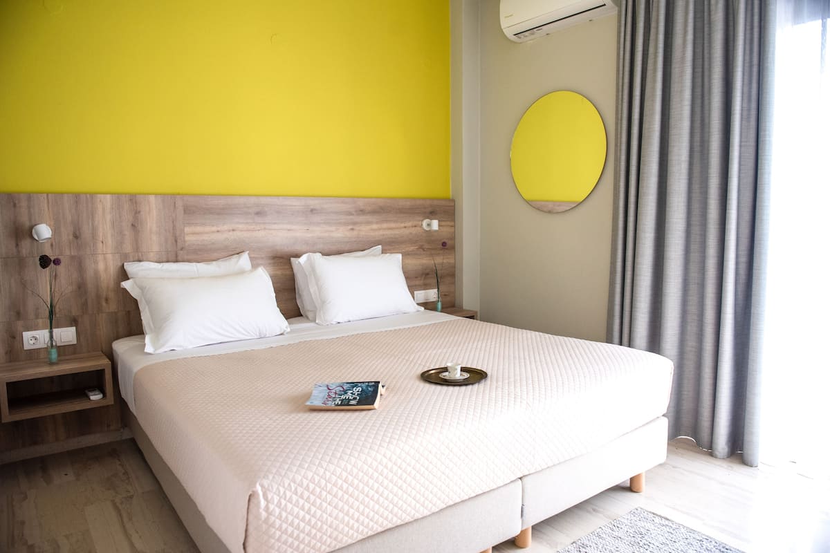 A neatly made bed with a white comforter and pillows, a bedside table with a book and a tray, and a round mirror on the wall.