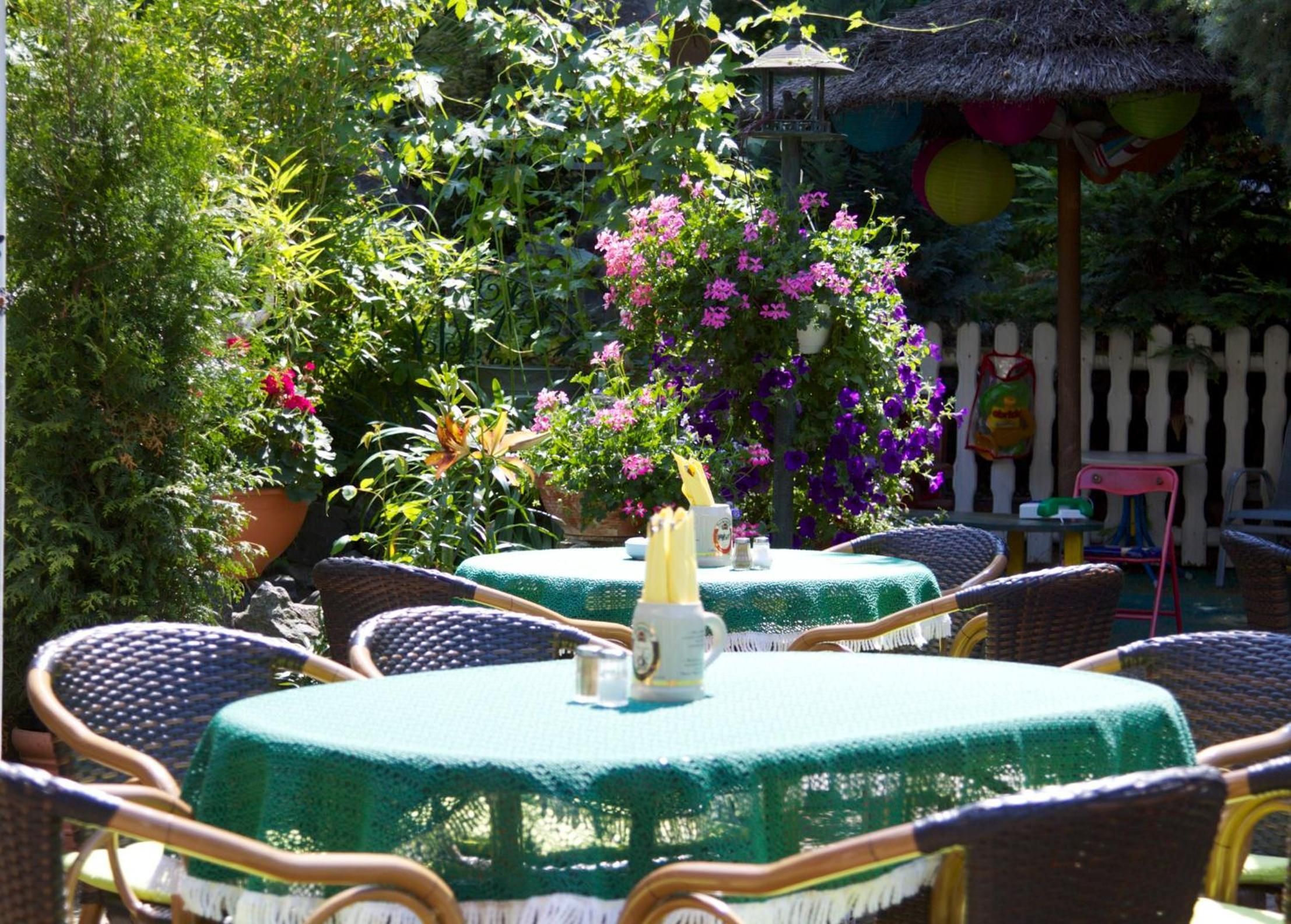 An outdoor seating area with round tables covered in green tablecloths, wicker chairs, and potted plants with pink and purple flowers.