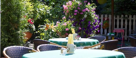 An outdoor seating area with round tables covered in green tablecloths, wicker chairs, and potted plants with pink and purple flowers.