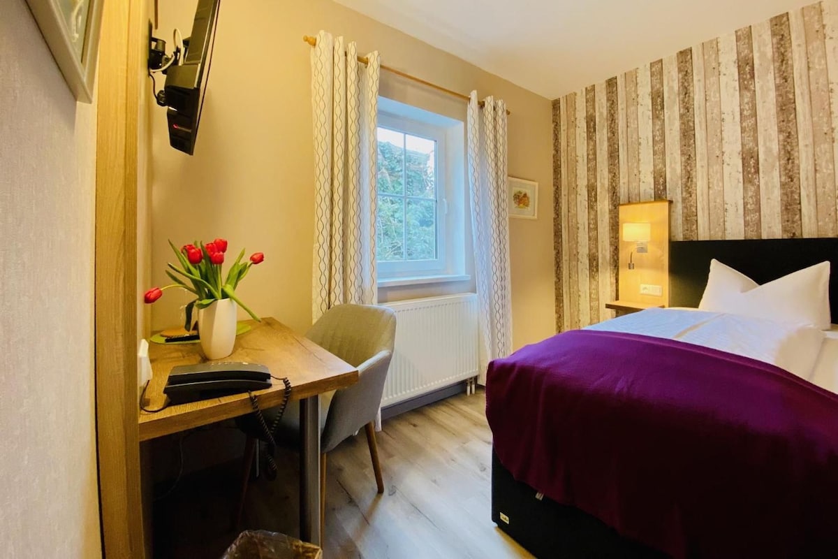 Habitación de hotel con cama, escritorio con teléfono, una silla, ventana con cortinas y pared con rayas verticales.