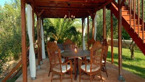 Un área de comedor al aire libre techada con una mesa redonda y sillas de mimbre, rodeada por una pérgola de madera y palmeras.