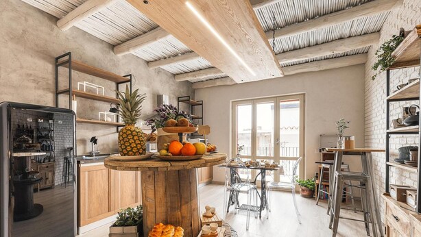 Una cocina moderna con vigas de madera, una isla central con fruta y pasteles, y un comedor con sillas de metal.