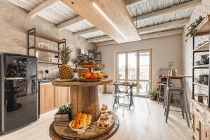 Una cocina moderna con vigas de madera, una isla central con fruta y pasteles, y un comedor con sillas de metal.