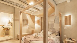 Un dormitorio con una cama grande, una mesita de noche, un banco de madera y un baño con lavabo y espejo.