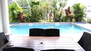 Area piscina con tavolo in legno e sedie in vimini, circondata da rigogliosa vegetazione tropicale.
