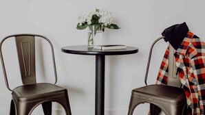Une petite table ronde sur laquelle est posé un vase de fleurs blanches, deux chaises en métal et une veste à carreaux jetée sur une chaise.