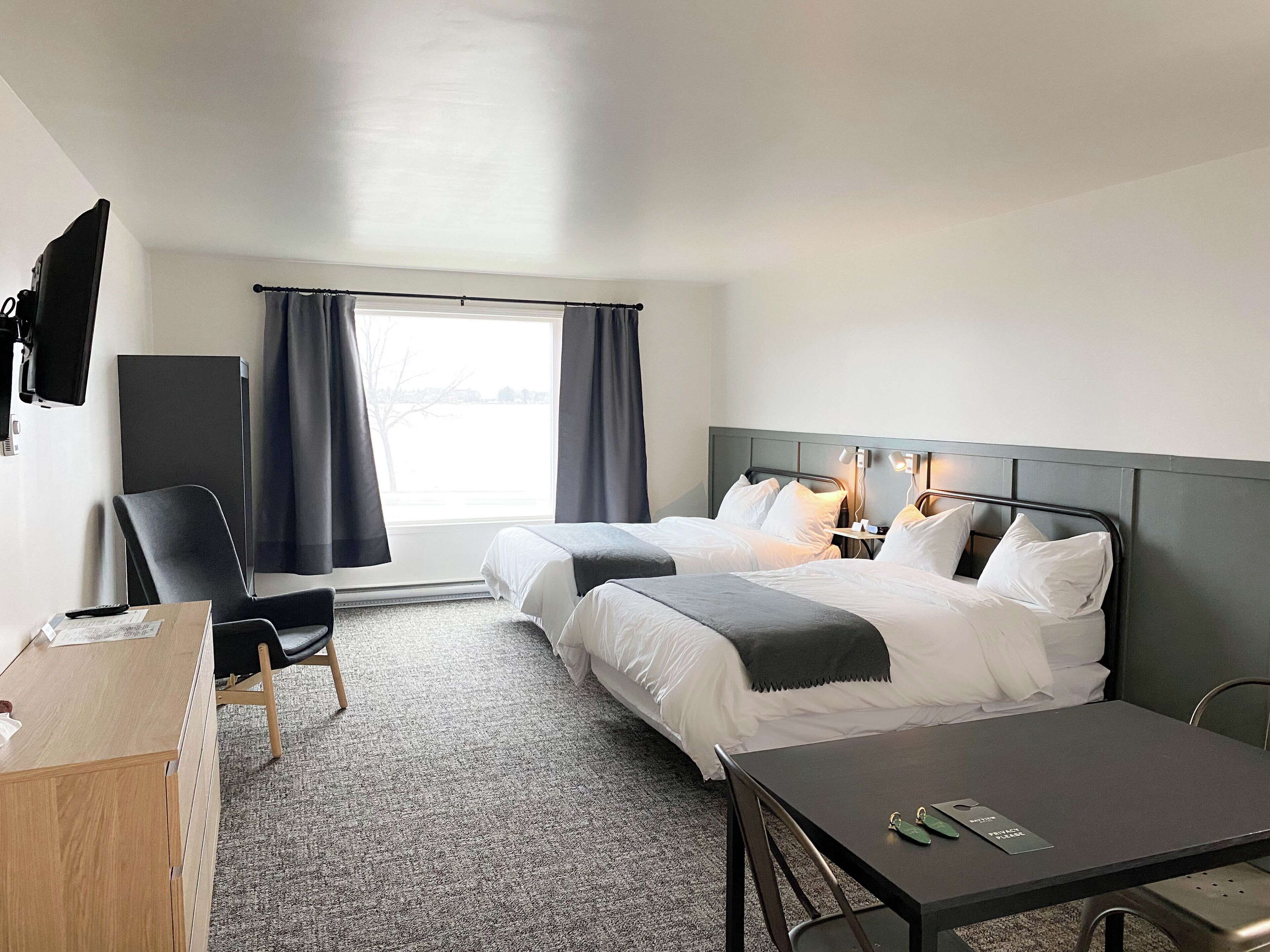 A hotel room with two beds, a desk, a chair, and a large window with curtains.