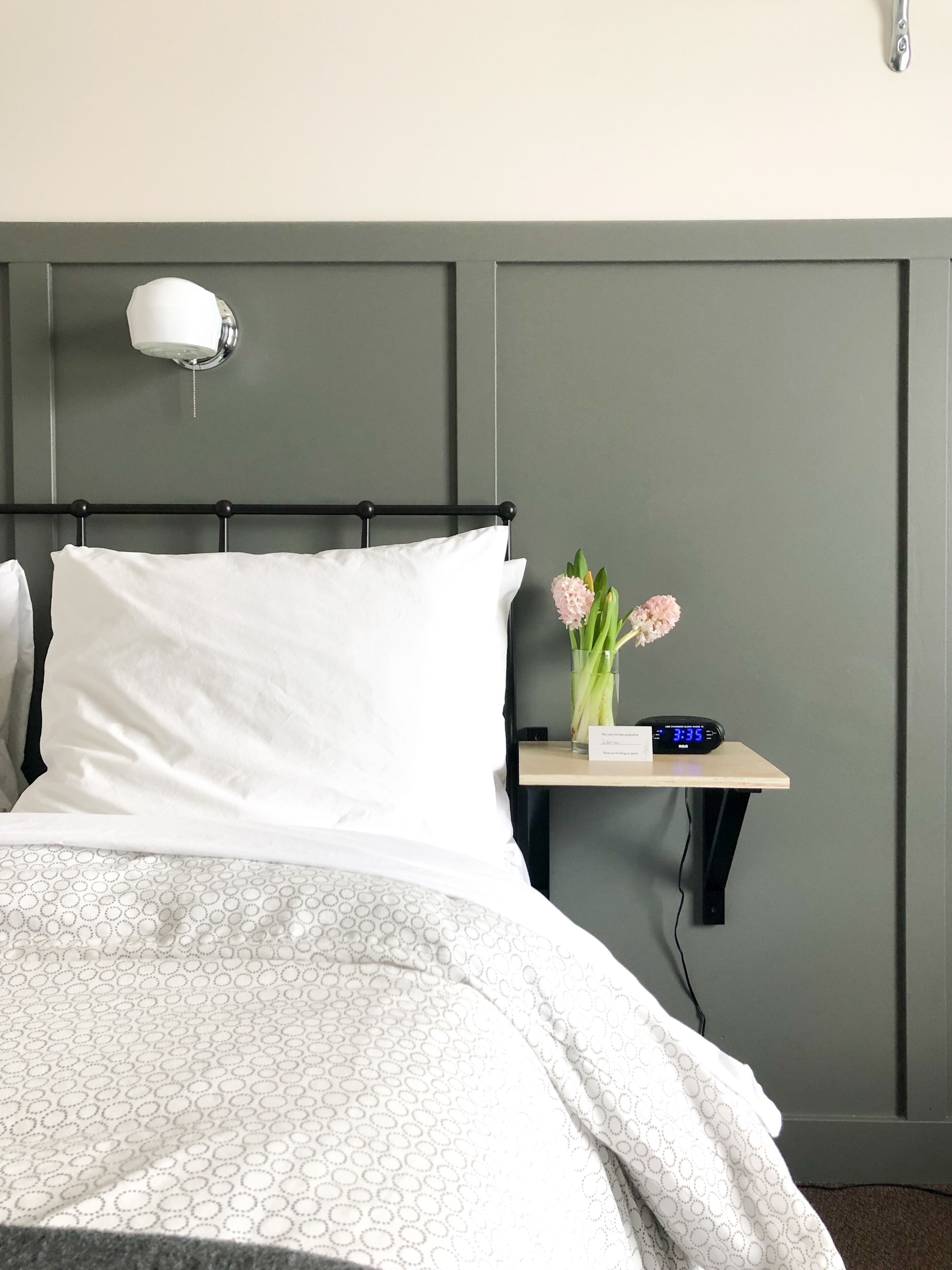 Una cama bien hecha con un edredón estampado, una mesita de noche con un reloj y un jarrón de flores, y una lámpara de pared.