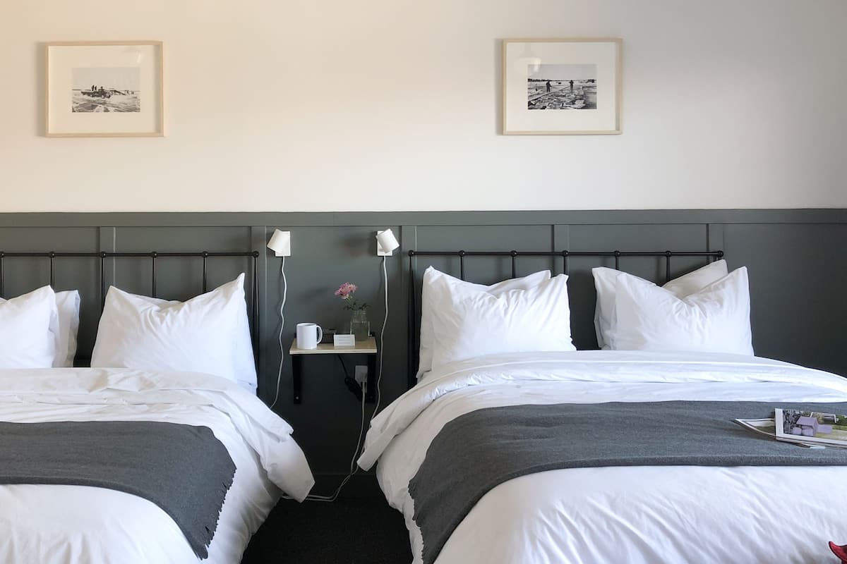 Quarto de hotel com duas camas, uma mesa de cabeceira e duas fotos emolduradas na parede.
