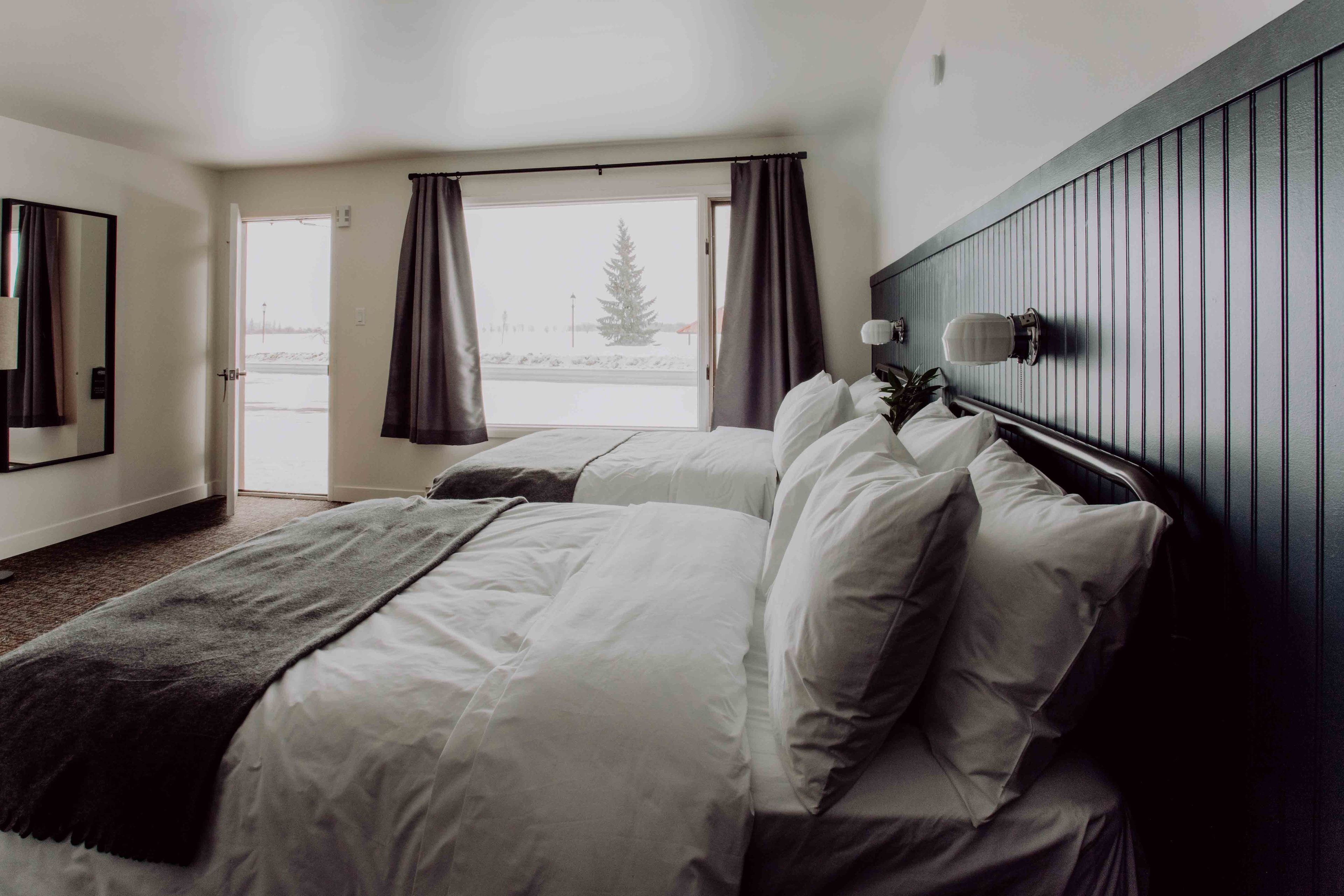 A hotel room with two beds, a large window with curtains, a wooden headboard, and a mirror on the wall.