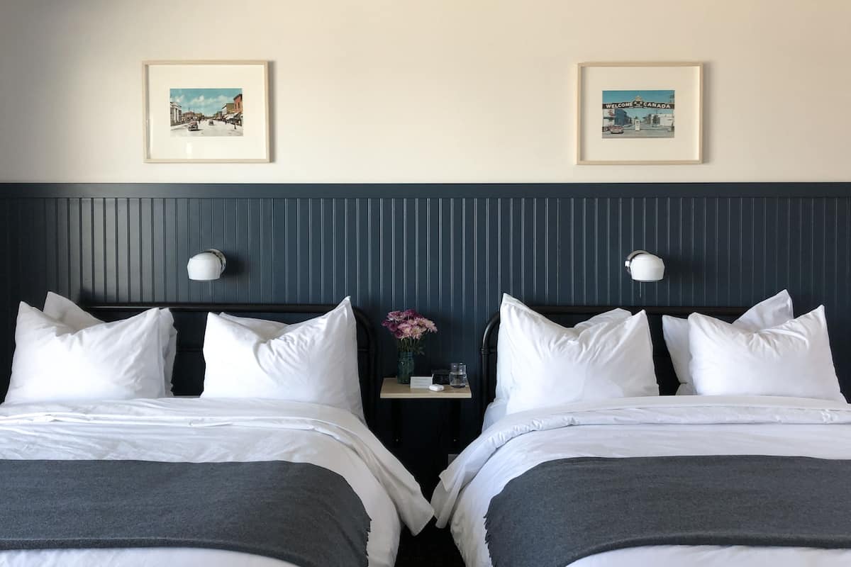 Quarto de hotel com duas camas, uma cabeceira escura e duas imagens emolduradas na parede.