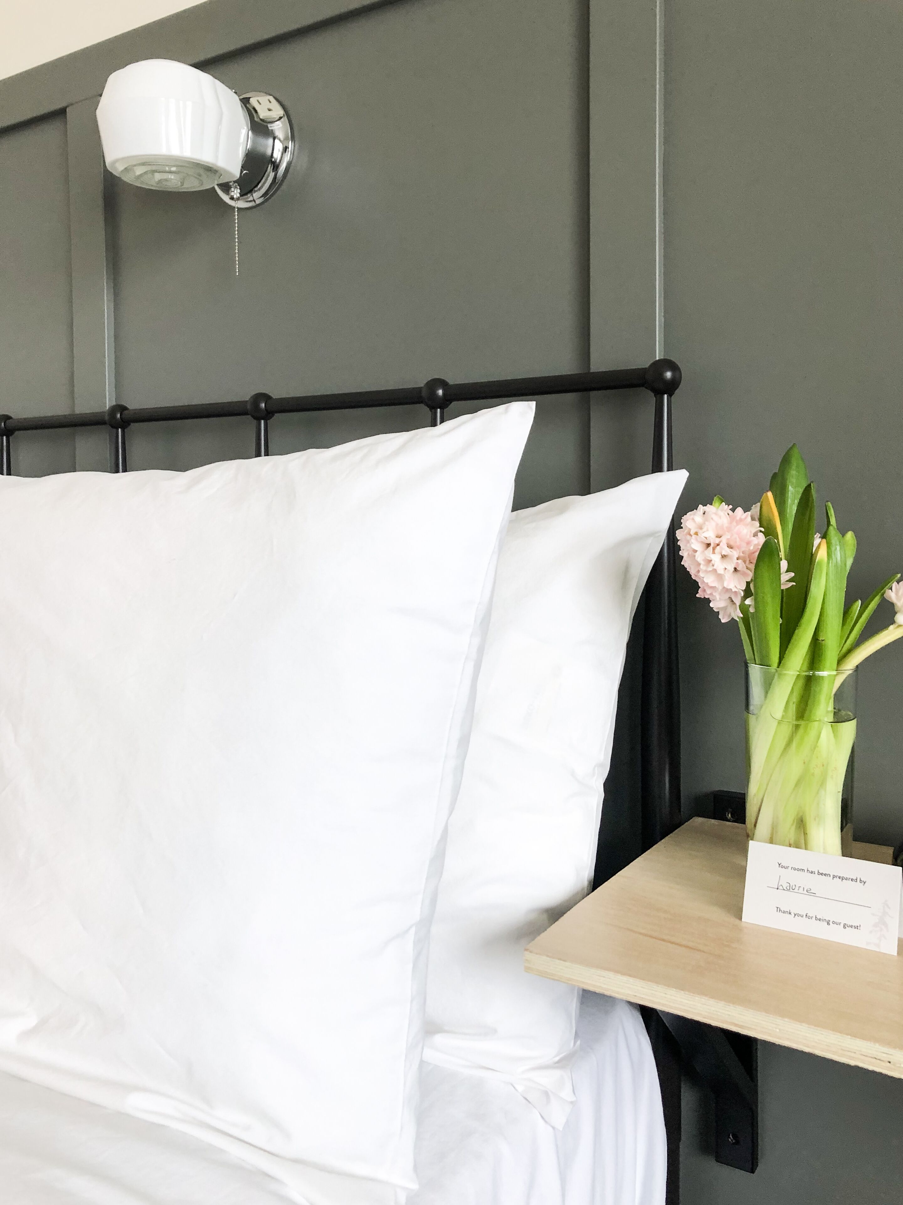A neatly made bed with white linens, a black headboard, and a bedside table with a vase of flowers and a thank you card.