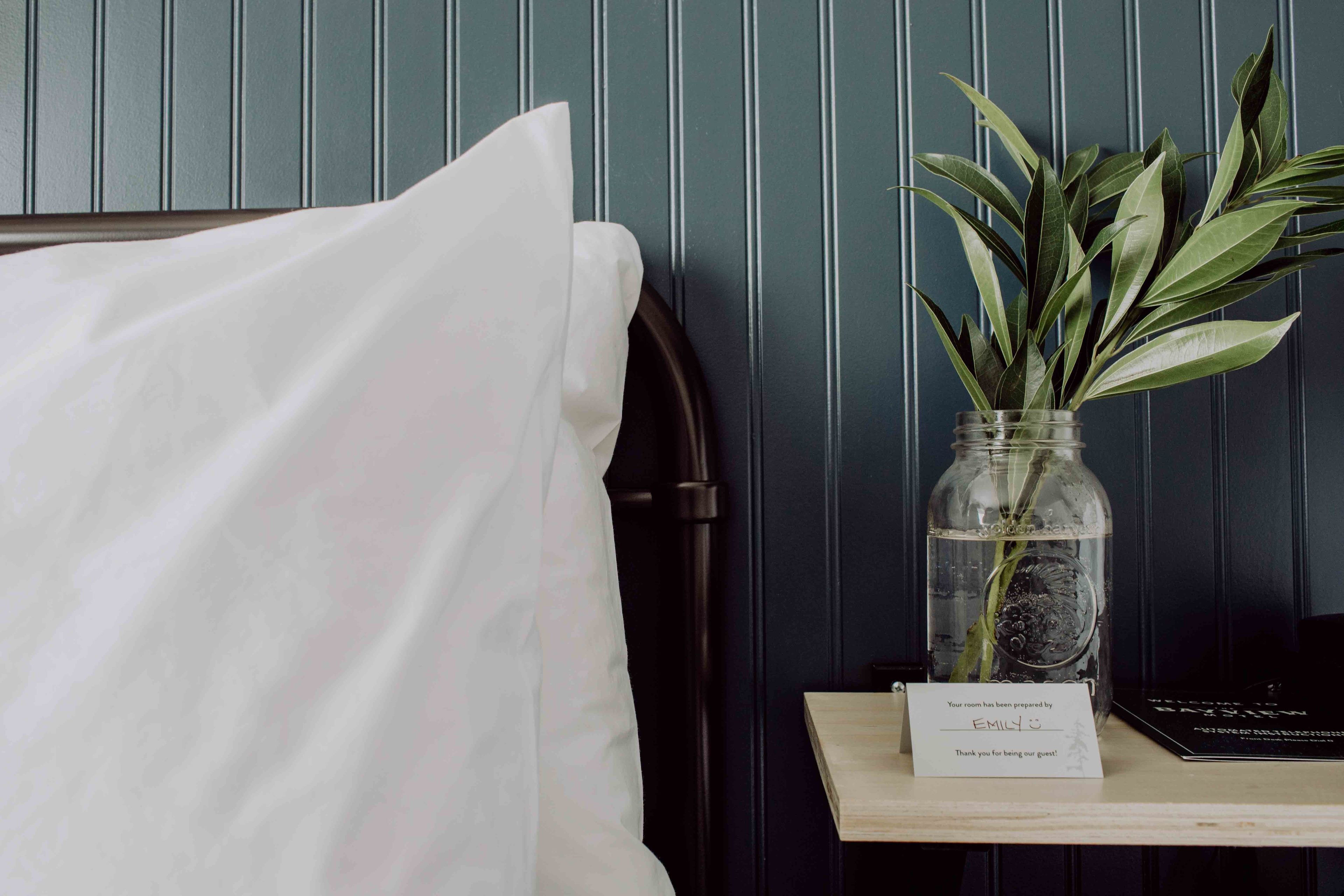 A neatly made bed with a white sheet, a small table with a vase of green leaves, and a sign indicating the room is prepared by Emily.