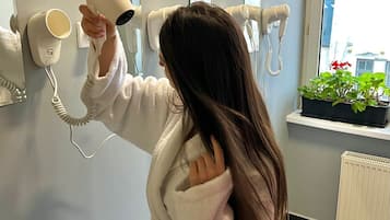 A person in a white robe using a hairdryer in a room with a window, a radiator, and a plant on the windowsill.