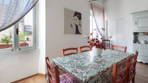 Uma sala de jantar com uma mesa posta para quatro pessoas, um vaso de flores, um quadro na parede e vista para o exterior através de uma janela.