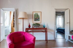 A room with a red chair, a lamp, a framed picture, and a small table with vases.
