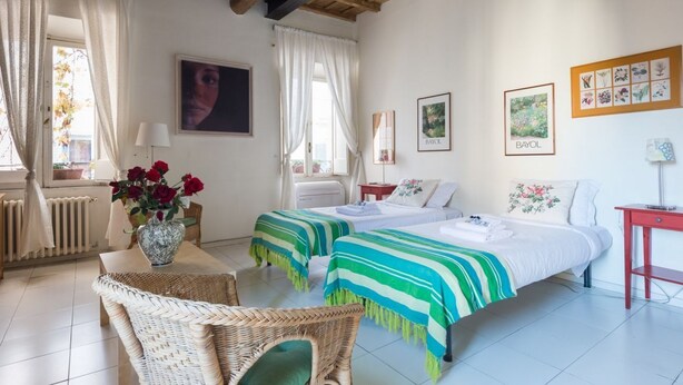 Quarto com duas camas, uma mesa com um vaso de flores, cadeiras de vime e uma mesinha lateral vermelha.