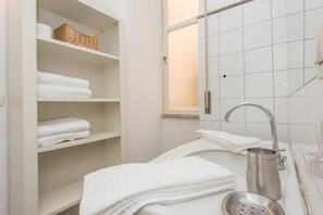 A bathroom with white tiled walls, a metal sink, a towel rack, and a shelf with folded towels. - Navona Charming Terrace Apartment (Rome)