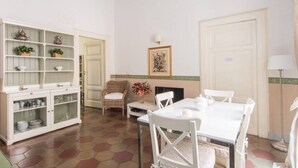 A dining area with a white table, chairs, a cabinet with glass doors, a wicker chair, a small table with a vase, and a painting on the wall. - Navona Charming Terrace Apartment (Rome)