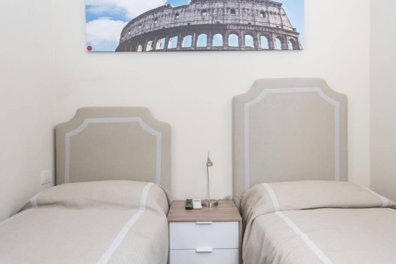 A hotel room with two beds, a nightstand with a lamp, and a picture of the Colosseum on the wall.