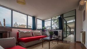 Ett modernt vardagsrum med en röd soffa, ett soffbord och stora fönster som ger utsikt över byggnader och träd.