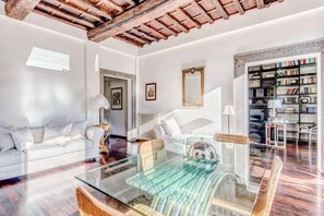 A spacious living room with a glass coffee table, white sofas, a wooden ceiling, and a bookshelf filled with books.