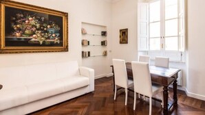 Una habitación con un sofá blanco, una mesa de comedor con sillas, una araña, una ventana con contraventanas blancas y un cuadro enmarcado en la pared.