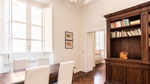 Una habitación con una mesa de comedor de madera, sillas blancas, una estantería y una puerta que da a otra habitación.