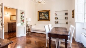 Un comedor con mesa y sillas de madera, un sofá blanco, una araña y una estantería llena de libros.