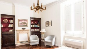 Una habitación con chimenea, dos sillones, una araña y una estantería.
