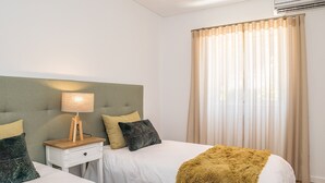 Una habitación de hotel con dos camas, una mesita de noche con lámpara, cabecera y ventana con cortinas.