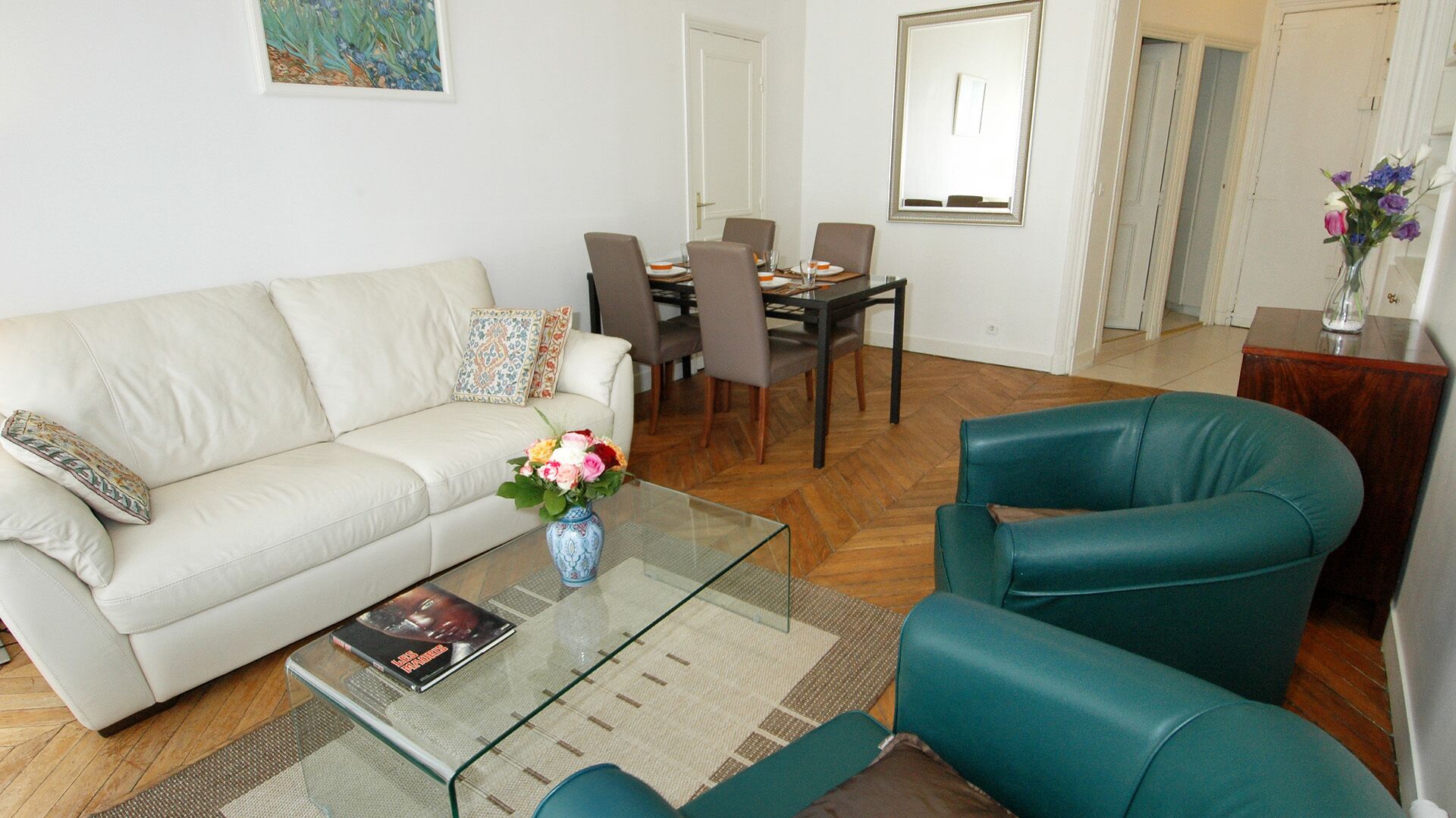 Ein Wohnzimmer mit einer Couch, einem Glastisch und einem blaugrünen Sessel. Im Hintergrund steht ein Esstisch mit Stühlen.