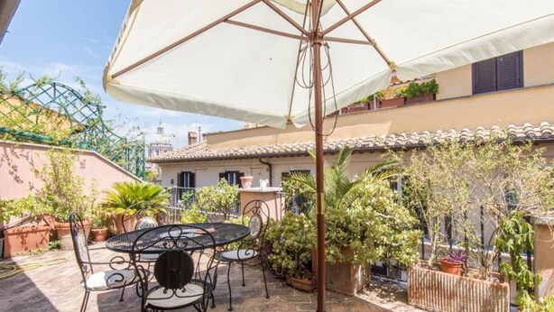 Un patio con una mesa y sillas, rodeado de macetas con plantas y un paraguas blanco.