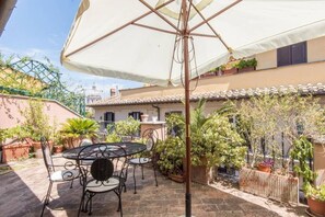 Eine Terrasse mit Tisch und Stühlen, umgeben von Kübelpflanzen und einem weißen Sonnenschirm.
