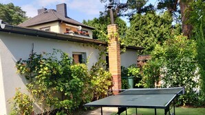 Une table de ping-pong installée dans un jardin, sur une pelouse artificielle, entourée de verdure, avec une maison en arrière-plan.