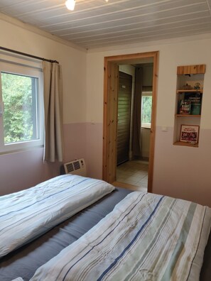 Quarto com cama, janela com cortinas, radiador, porta que dá para outro cômodo e prateleira com livros.