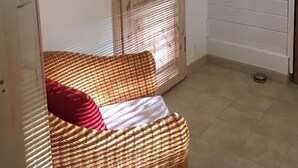 Une pièce avec une porte en bois, un tableau encadré au mur, un canapé en osier avec un coussin rouge et un mur blanc avec un câble rouge.