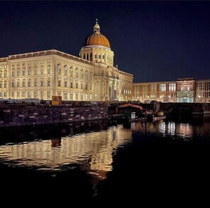 Um edifício grande e histórico com uma cúpula imponente e arquitetura clássica, localizado às margens de um corpo d'água calmo que reflete a estrutura.