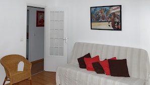 Una sala de estar con un sofá blanco, cojines rojo y marrón, un sillón de mimbre y una puerta con un panel de vidrio.