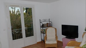 Una sala de estar con televisor, sillas de mimbre, una estantería y una puerta que da a un balcón.