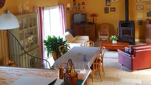 A spacious living area with a high ceiling, wooden beams, a dining table, chairs, a sofa, a TV, and a wood-burning stove. - Charming cottage in the country of Futuroscope (Persac)