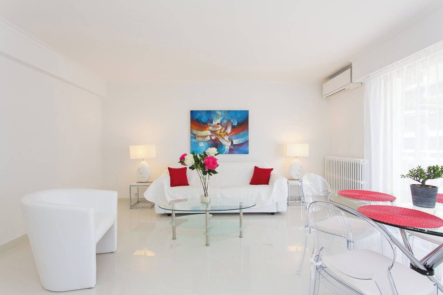 Una sala de estar moderna con una decoración blanca, una mesa de centro de vidrio, un sofá con cojines rojos y un cuadro en la pared.